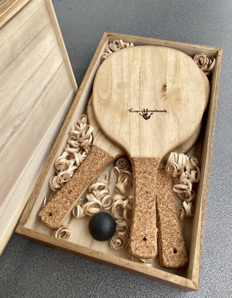 Handmade Frescobol paddles crafted from sustainable paulownia wood, presented in a natural wooden box with decorative shavings and a game ball