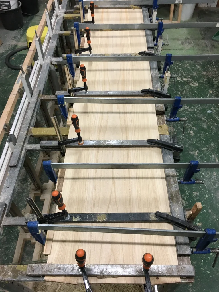 Paulownia wood panels being laminated and pressed with clamps in a workshop. The fine grain and lightweight properties make it ideal for surfboard construction.